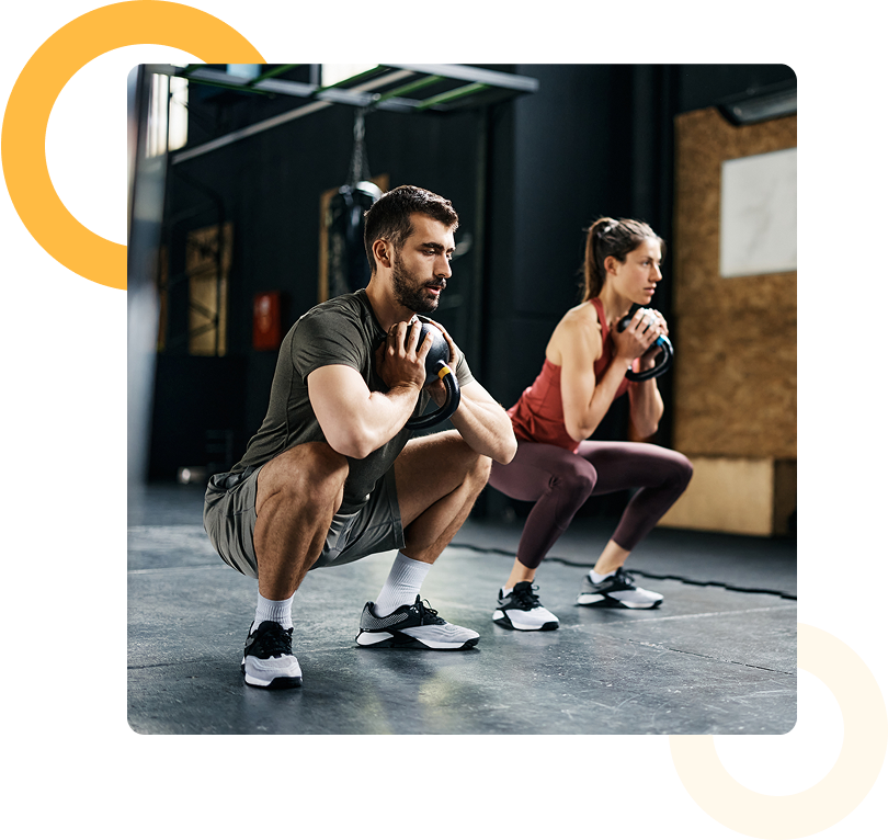 A man and a woman perform squats while holding weights at chest level in a gym setting.