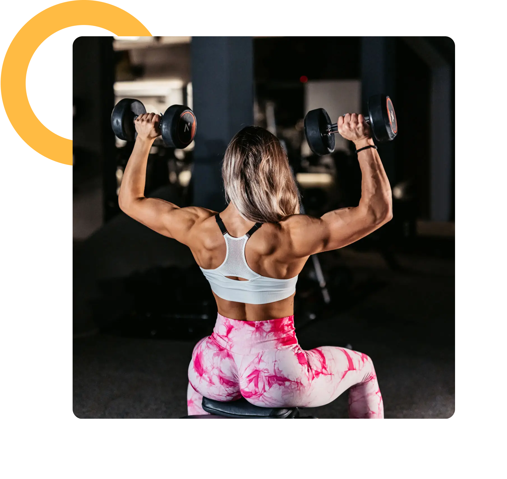 A woman in a gym performs a shoulder press with dumbbells, seated on a bench, wearing a white sports bra and pink patterned leggings.