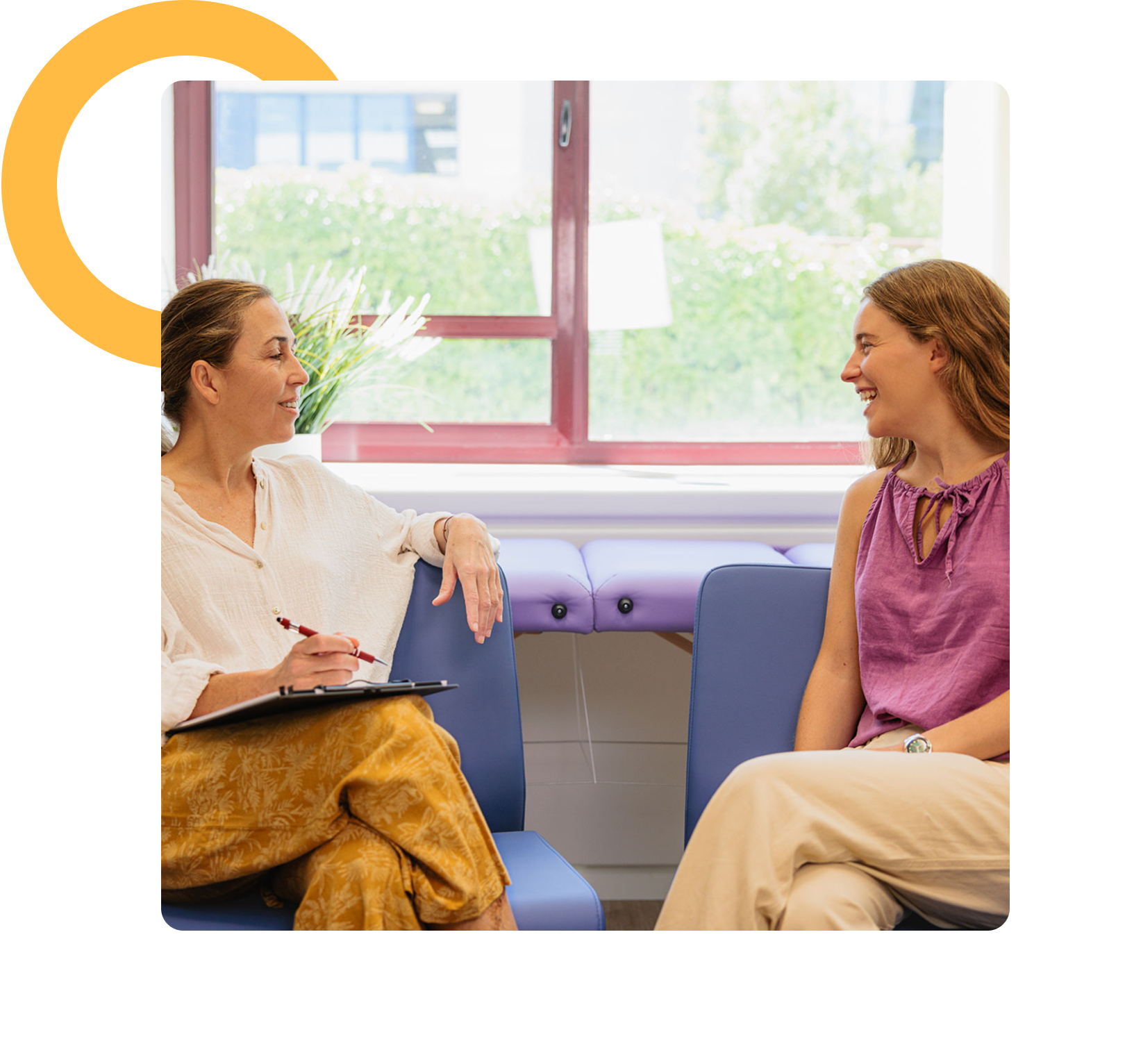 Two women sit on blue chairs in a bright room, having a conversation. One holds a clipboard and pen, while the other smiles, suggesting a counseling or interview setting.