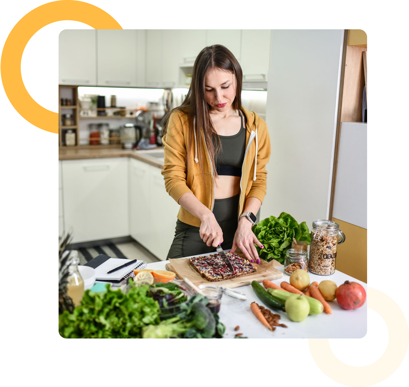 Woman in a kitchen slicing a granola bar on a cutting board, surrounded by fresh vegetables, fruit, and jars of grains.
