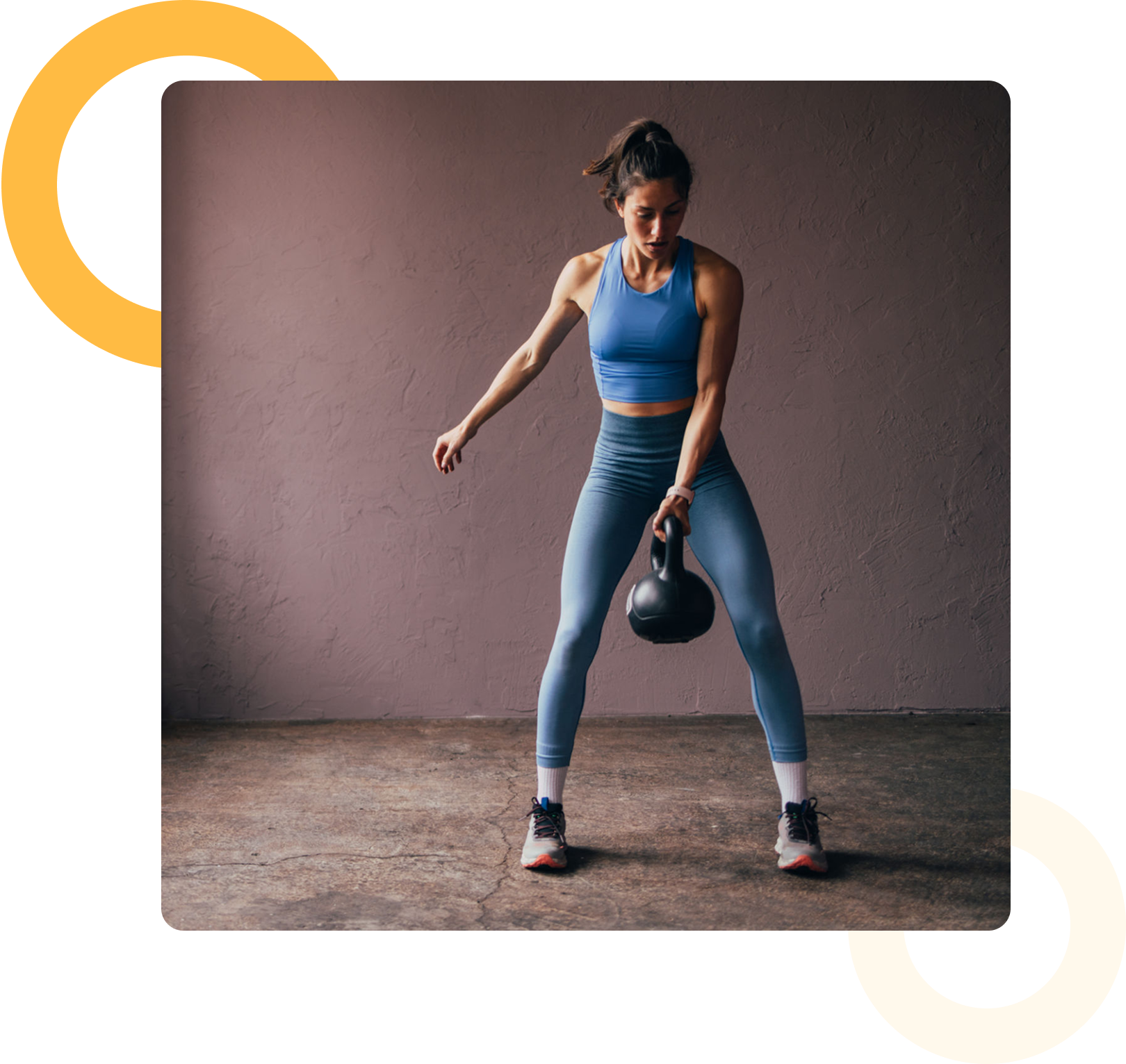 A woman in athletic wear swings a kettlebell with one hand in a minimalist indoor space with a concrete floor and plain wall.