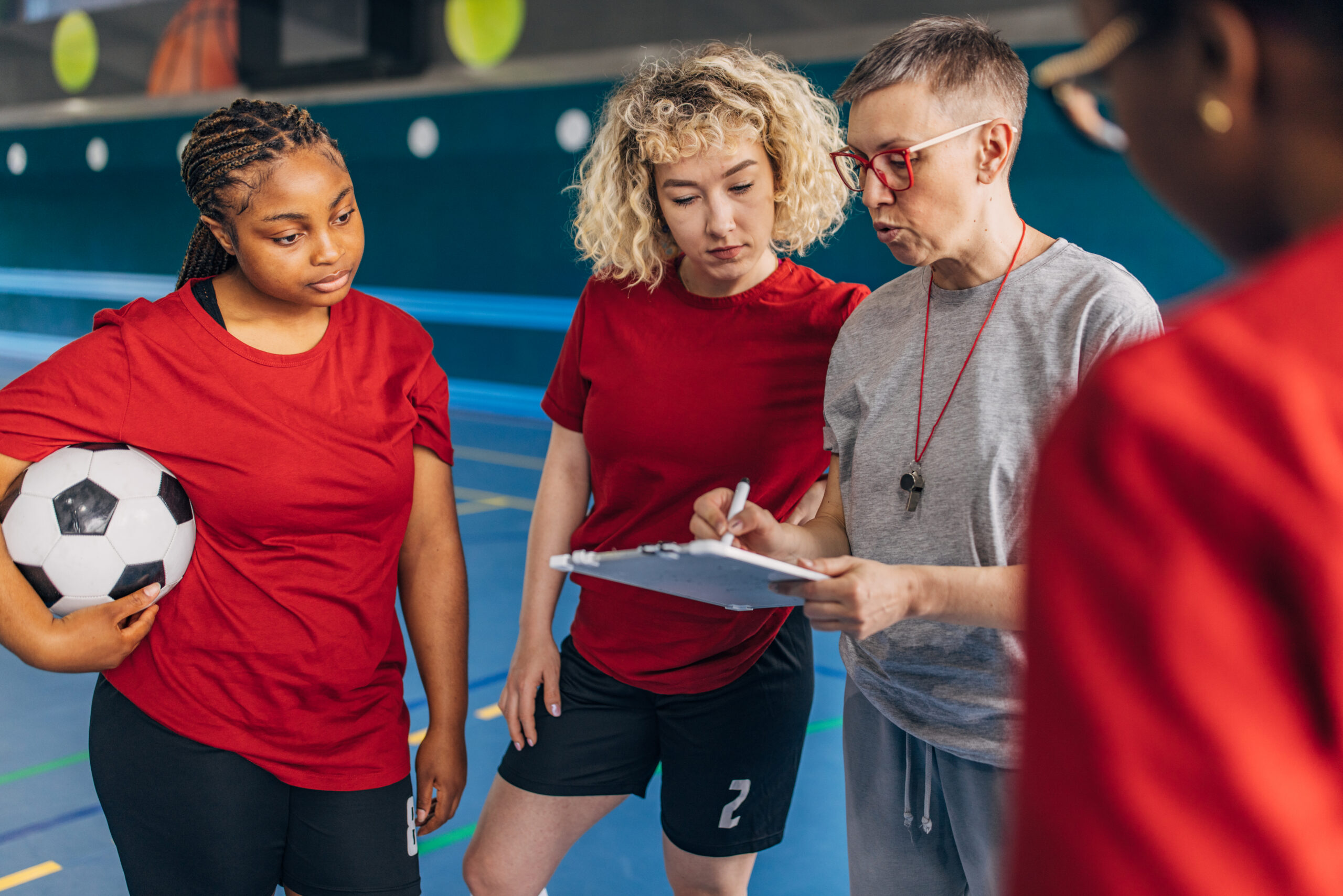 A soccer coach is instructing a group of attentive female players on an indoor court. The players, in red shirts, focus on the coach's instructions and one holds a soccer ball.