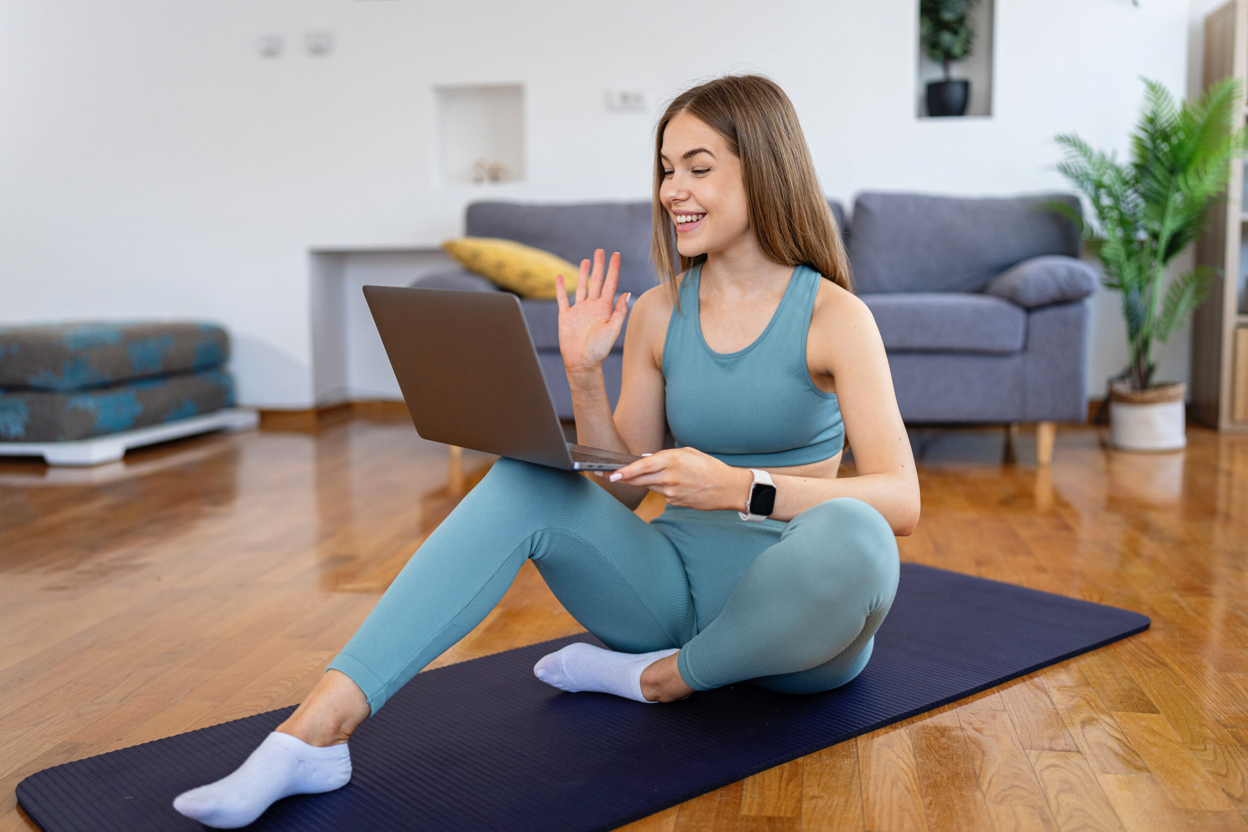 Side view of a happy smiling young woman in blue sportswear sitting on the floor, waving at her laptop screen during an online fitness class in a bright living room.
