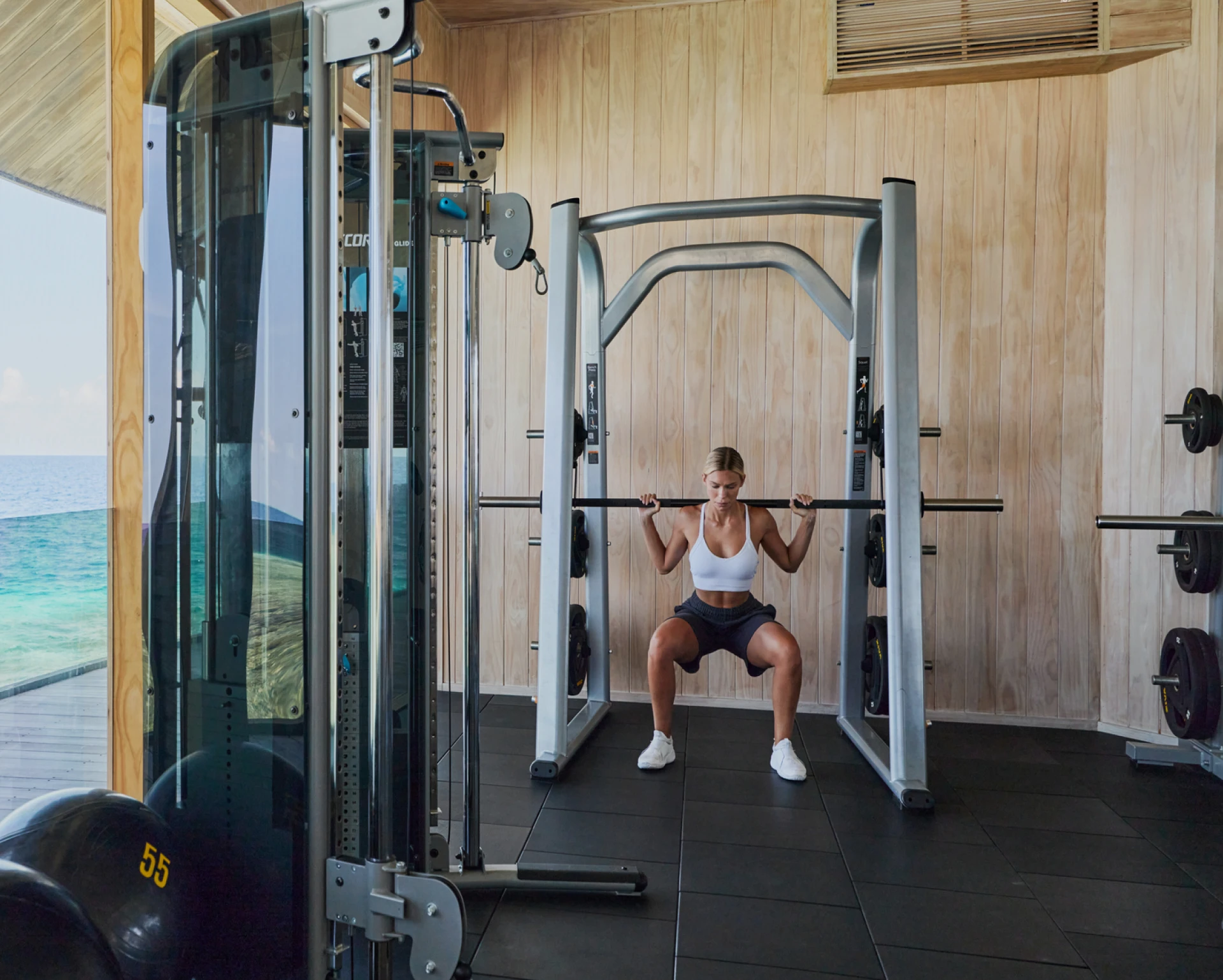 A person performs a squat exercise with a barbell in a gym with wooden walls and various fitness equipment visible.