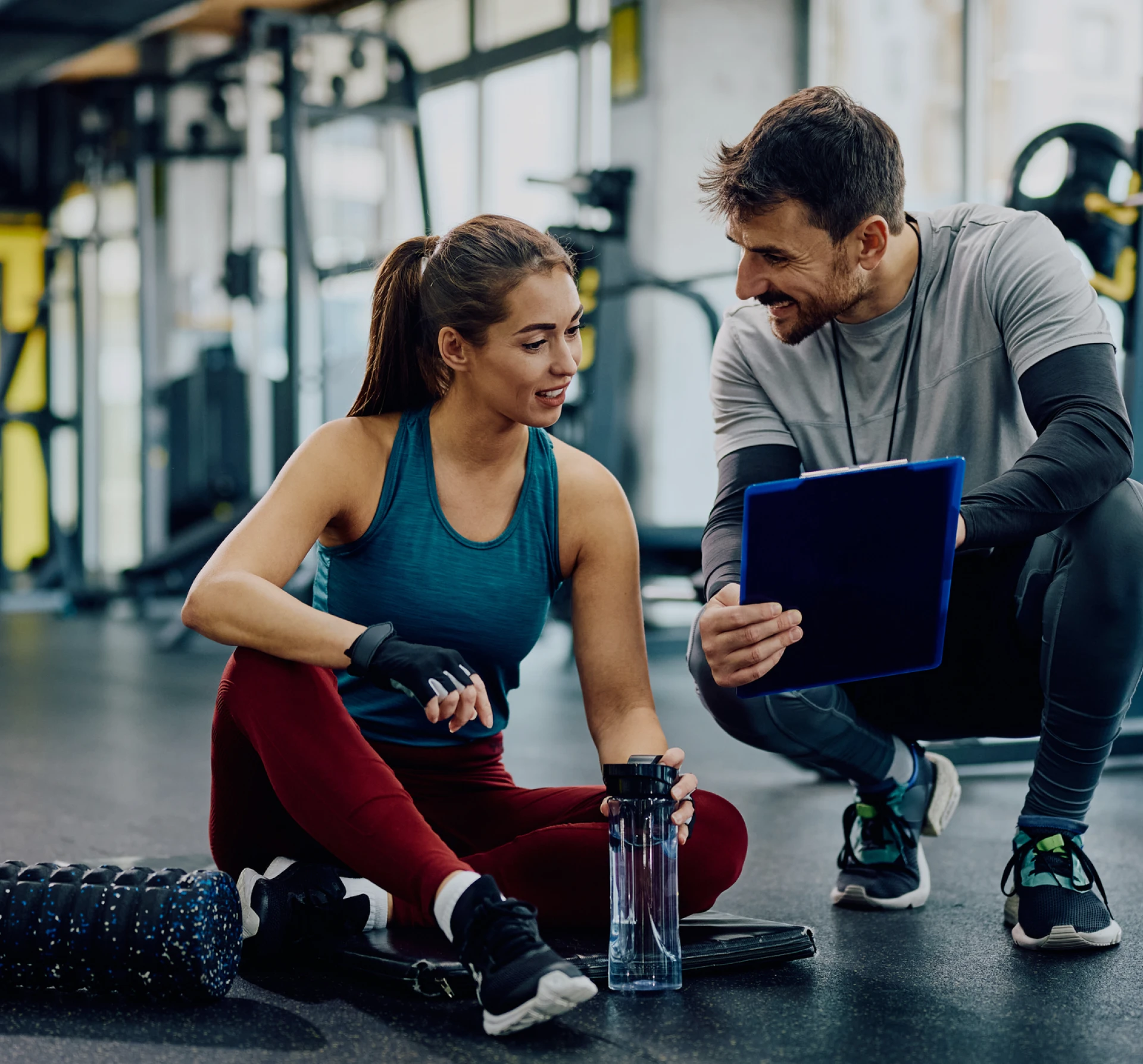 A woman sits on the gym floor holding a water bottle, while a man kneels beside her showing her information on a clipboard. Gym equipment is visible in the background.