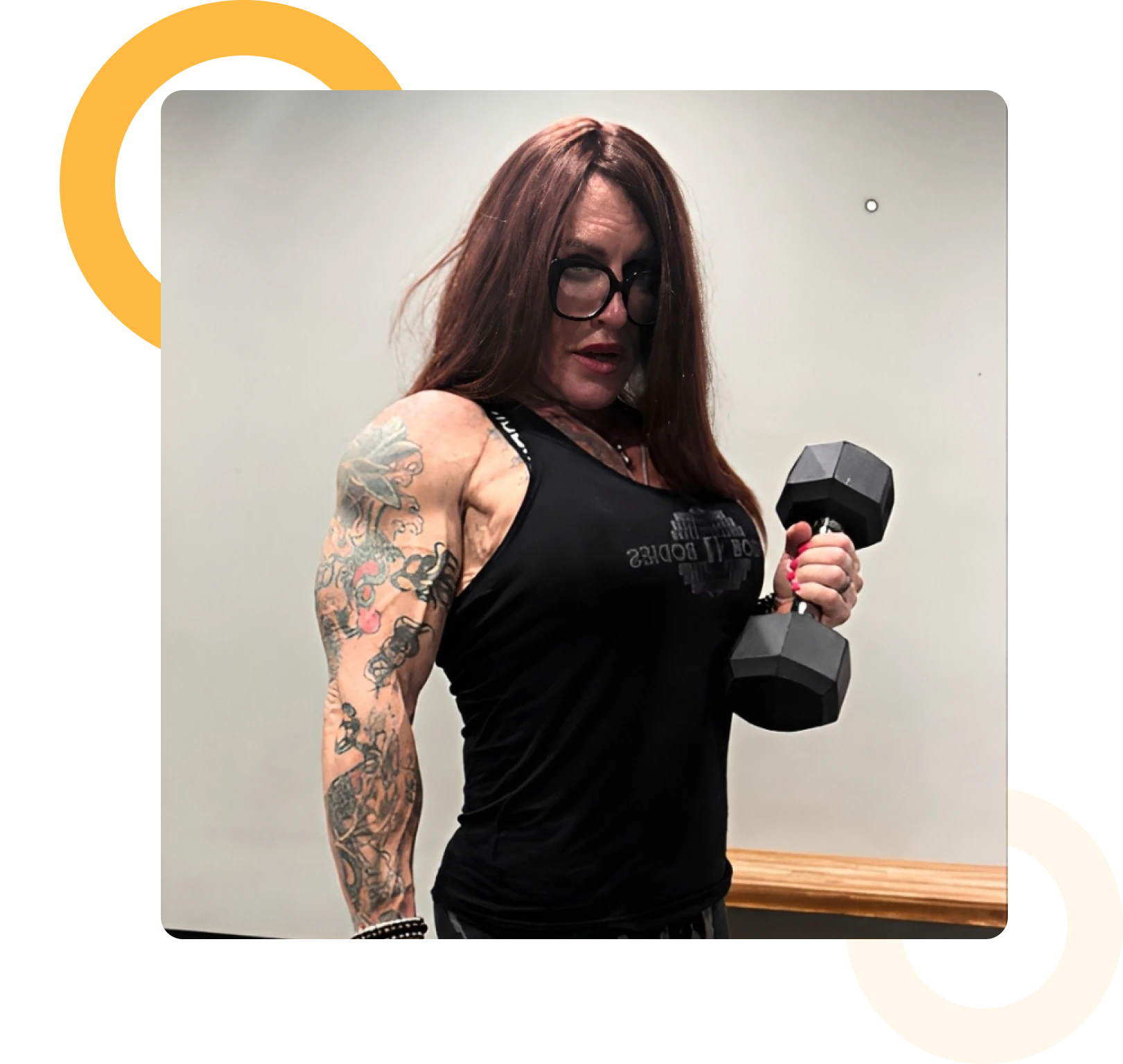Person with long brown hair and glasses holding a dumbbell, wearing a sleeveless black shirt, shows tattooed arm in a gym setting.