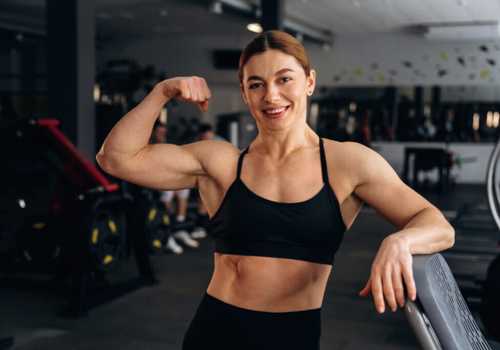Woman athlete shows off her muscular bicep while smiling in a gym, with various exercise machines and weights visible in the background