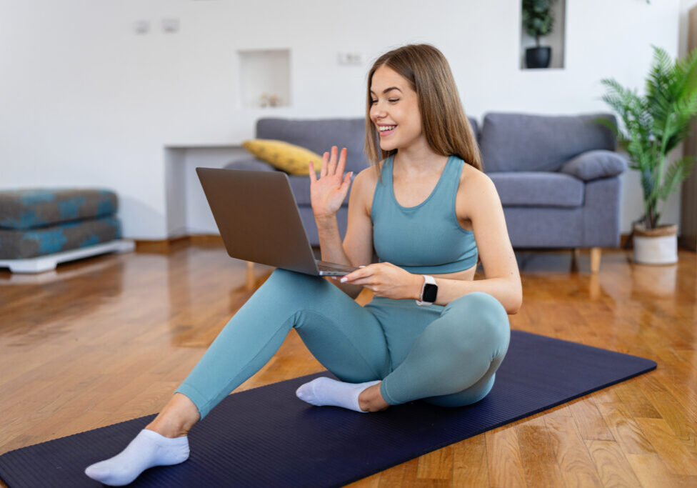 Side view of a happy smiling young woman in blue sportswear sitting on the floor, waving at her laptop screen during an online fitness class in a bright living room.